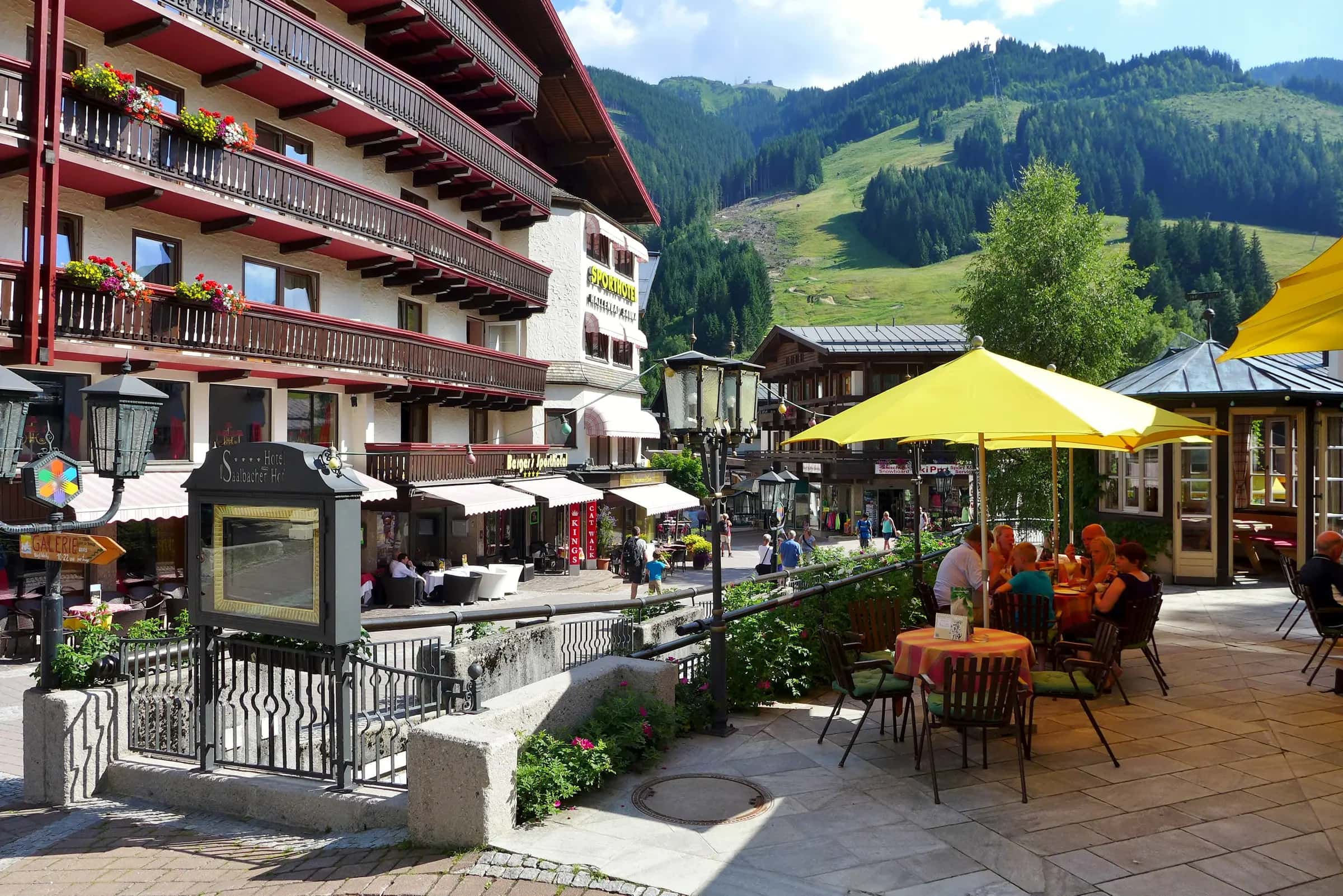 View of Dorfplatz (village square) and several hotels in Saalbach, a village of Saalbach-Hinterglemm municipality, Salzburg (state), Austria. In the background is Schattberg mountain; the first section of its cableway is visible at centre right, and the upper terminus is barely visible at top centre.