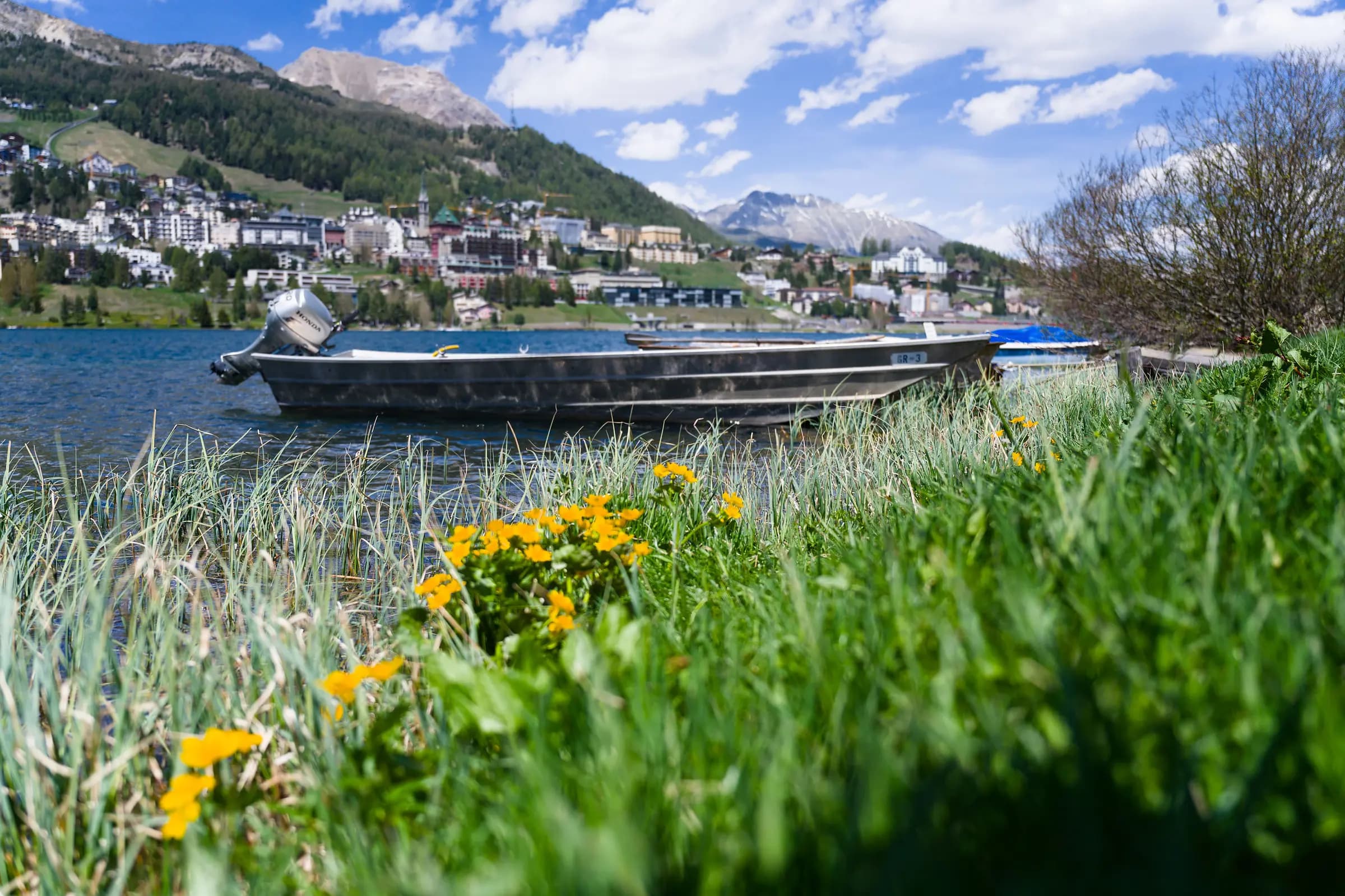 A boat rests by Lake St. Moritz with vibrant flowers and a stunning mountain backdrop.