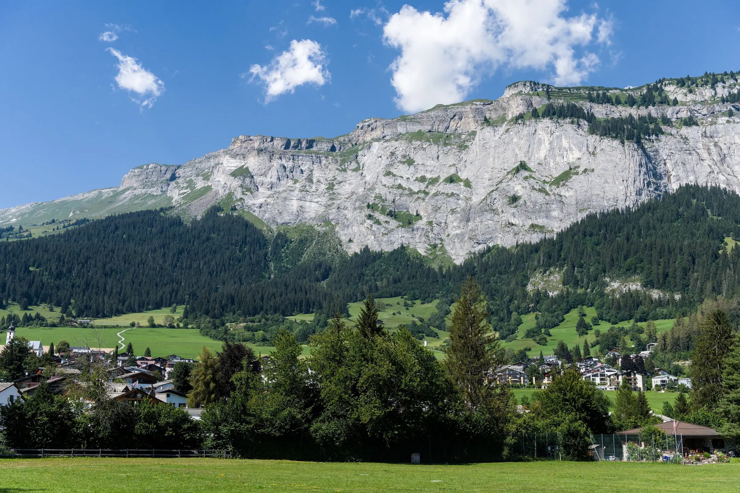 Stunning view of alpine village with majestic mountains and lush greenery under a bright blue sky.