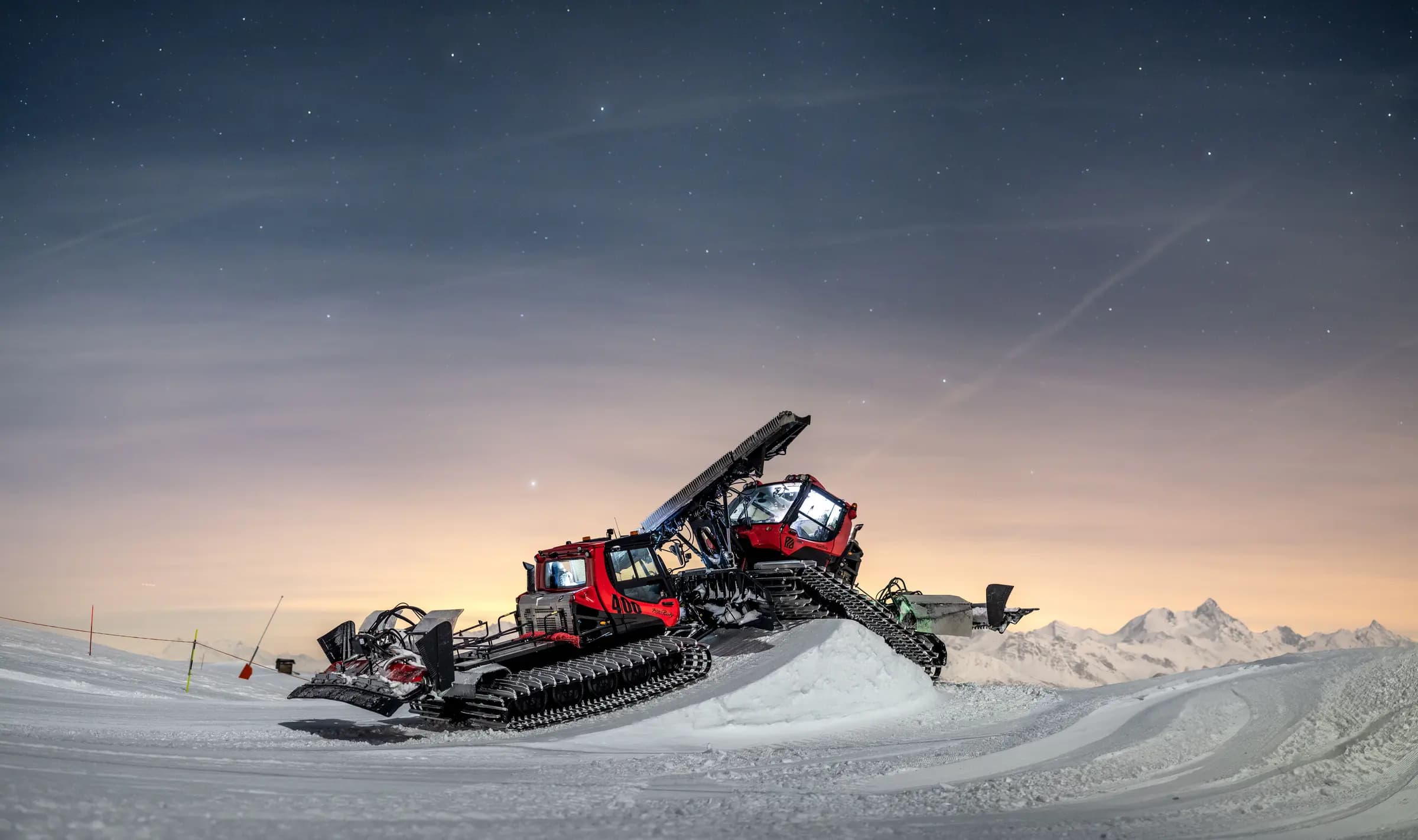 Night scene of snow grooming machinery in the Swiss Alps under a star-filled sky, surrounded by snowy mountains.