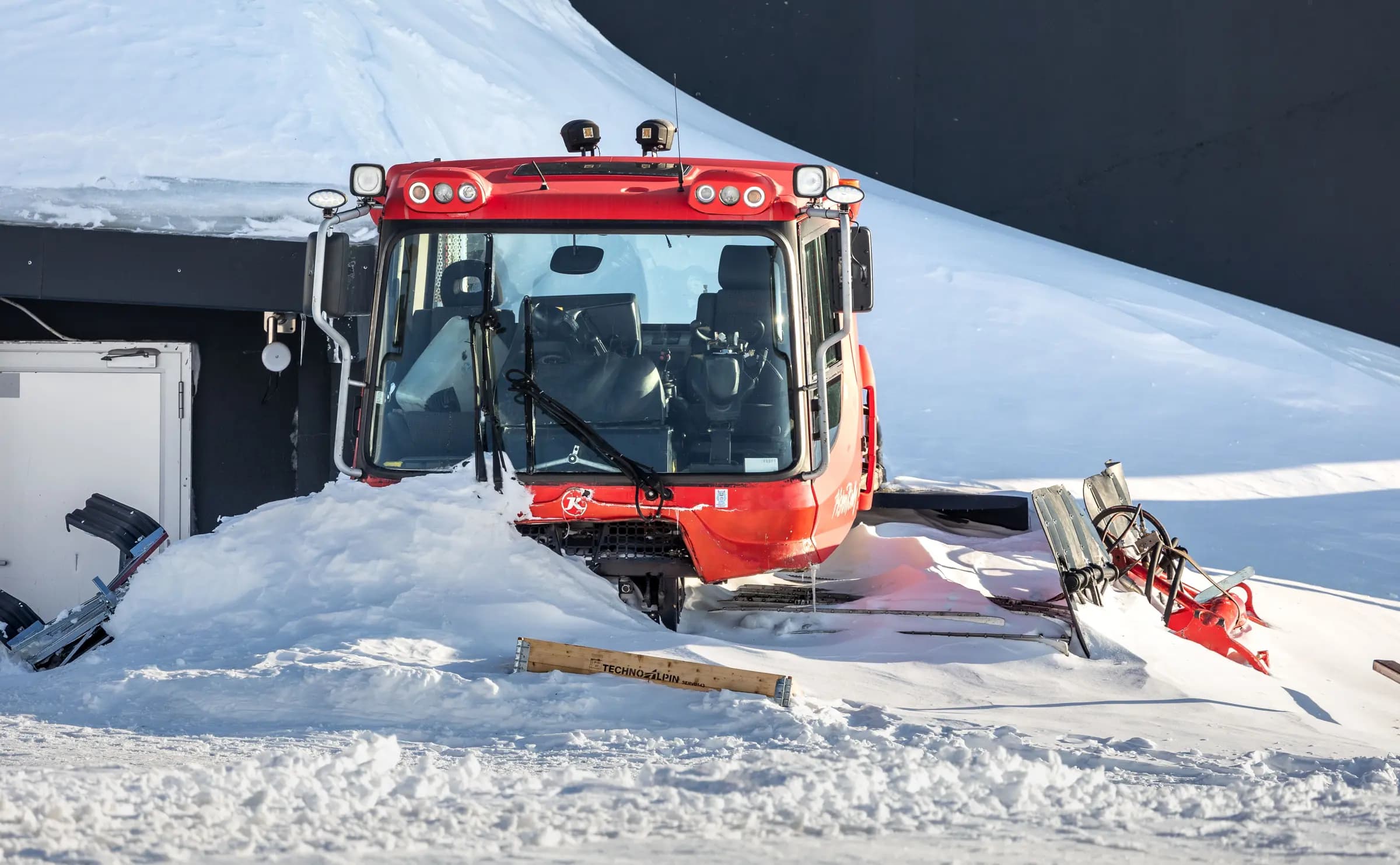 A red snow groomer partially buried in snow on a ski slope, ready for maintenance.