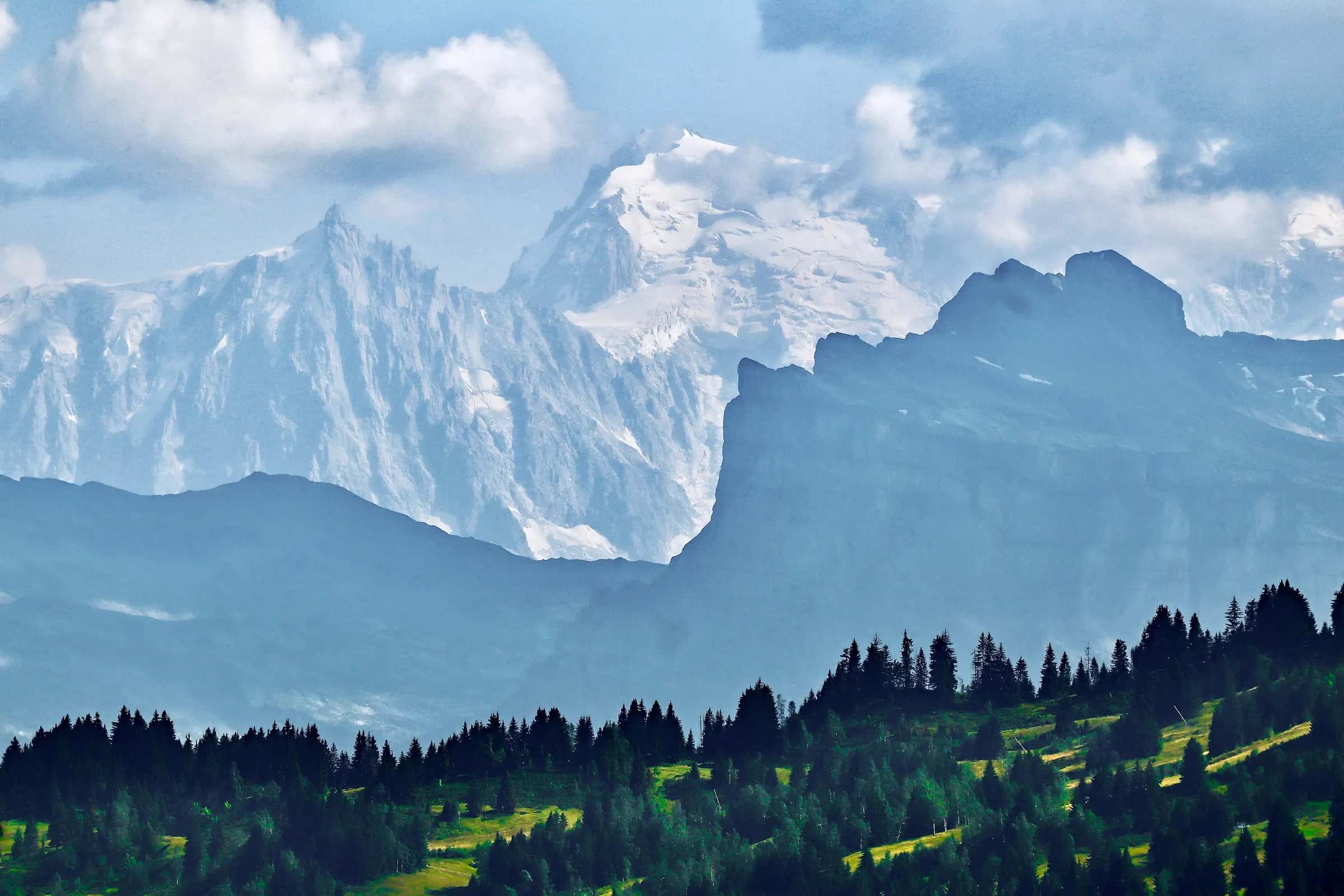 I took this photo during summer from the ski lift at Secteur Mont Chéry ski resort (elevation approximately 1700m), above the village of Les Gets. The intermediate black mountain with the precipice is Tête à l'Âne of 2800m elevation. In the background, the jagged rocky peak on the left is Aiguille du Midi (about 3850m), while the big snowy mountain is Mont Blanc du Tacul (about 4250m high). The main peak of Mont Blanc is off to the right, shrouded in cloud.