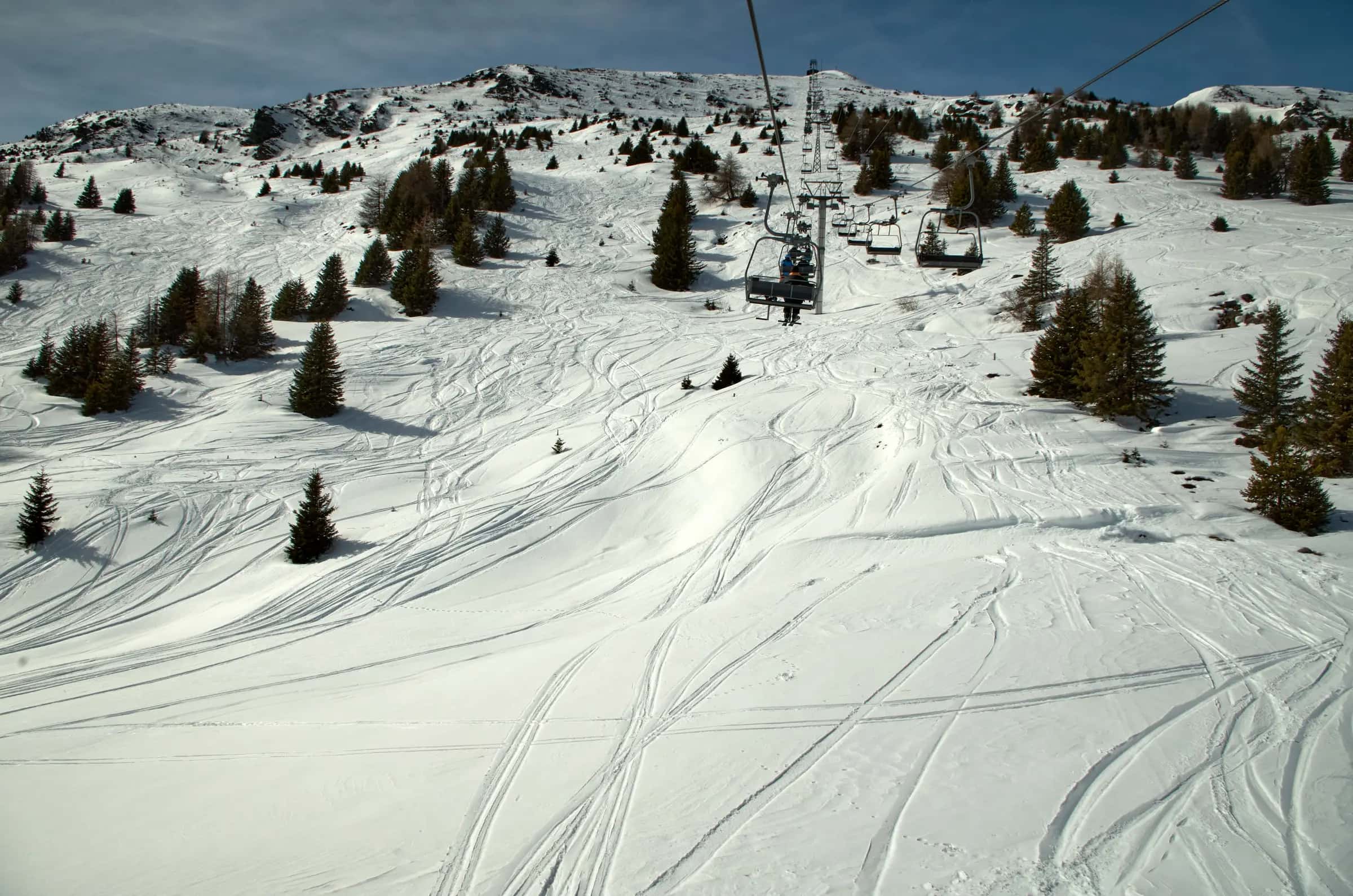 A picturesque ski slope with chairlift, snow tracks, and pine trees under a clear winter sky.